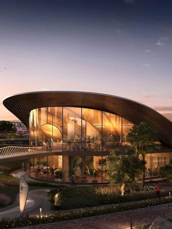 Futuristic building with a curved roof and glass walls, surrounded by greenery, under a twilight sky. Pathways and lights accentuate the scene.