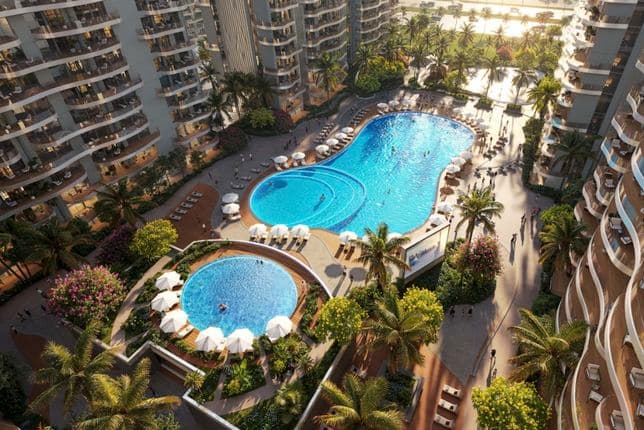 An aerial view of a tropical resort with two blue pools, palm trees, white umbrellas, lounge chairs, and curved high-rise buildings.