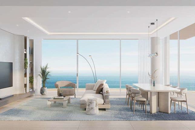 Spacious modern living and dining area with floor-to-ceiling windows overlooking the ocean, featuring a light neutral sofa, minimalist furniture, a large blue gradient rug, and an open balcony with coastal views.
