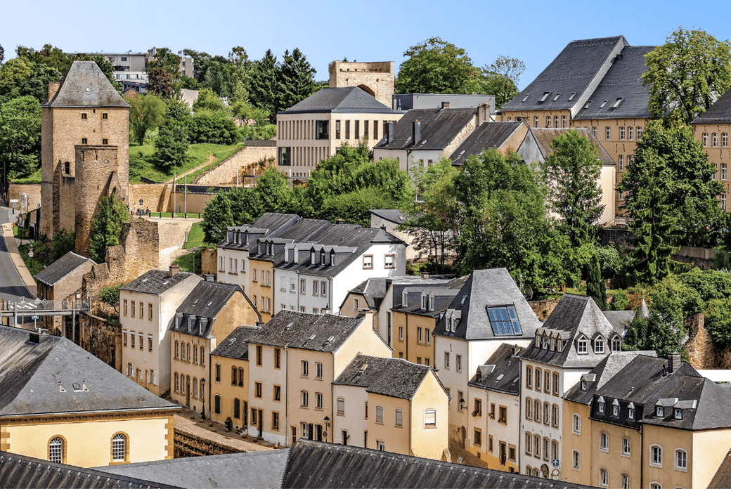 Luxembourg city view with stone buildings, tower, and green trees under a blue sky.
