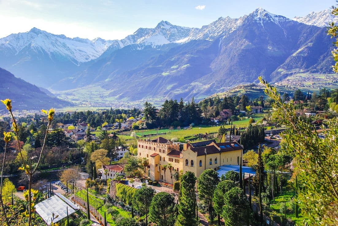 Eine malerische Aussicht auf eine gelbe Villa, umgeben von üppigem Grün, mit schneebedeckten Bergen im Hintergrund unter einem klaren blauen Himmel.