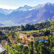 Eine malerische Aussicht auf eine gelbe Villa, umgeben von üppigem Grün, mit schneebedeckten Bergen im Hintergrund unter einem klaren blauen Himmel.