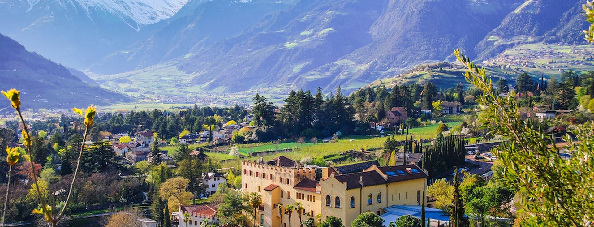Eine malerische Aussicht auf eine gelbe Villa, umgeben von üppigem Grün, mit schneebedeckten Bergen im Hintergrund unter einem klaren blauen Himmel.