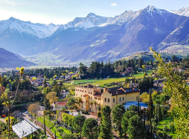 Eine malerische Aussicht auf eine gelbe Villa, umgeben von üppigem Grün, mit schneebedeckten Bergen im Hintergrund unter einem klaren blauen Himmel.