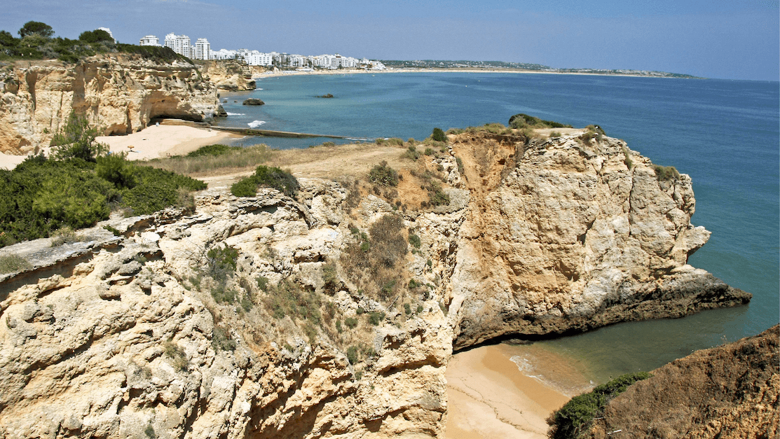 Rocky cliffs overlooking a sandy beach with turquoise waters, distant white buildings, and a clear blue sky.