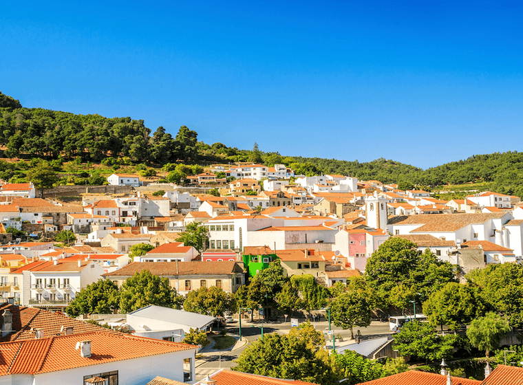 Landschaftsblick auf eine malerische Stadt mit bunten Häusern und roten Ziegeldächern vor einem üppigen, grünen Hügel unter einem klaren blauen Himmel.
