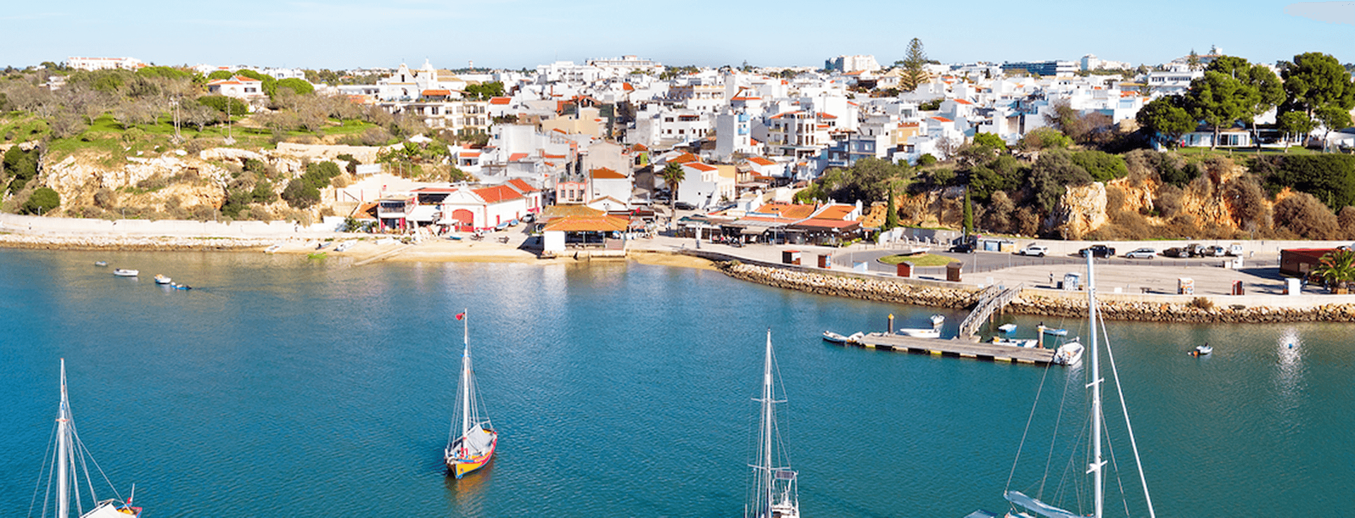 Vista panorâmica de uma vila costeira com edifícios brancos, veleiros sobre águas calmas e um céu azul límpido.