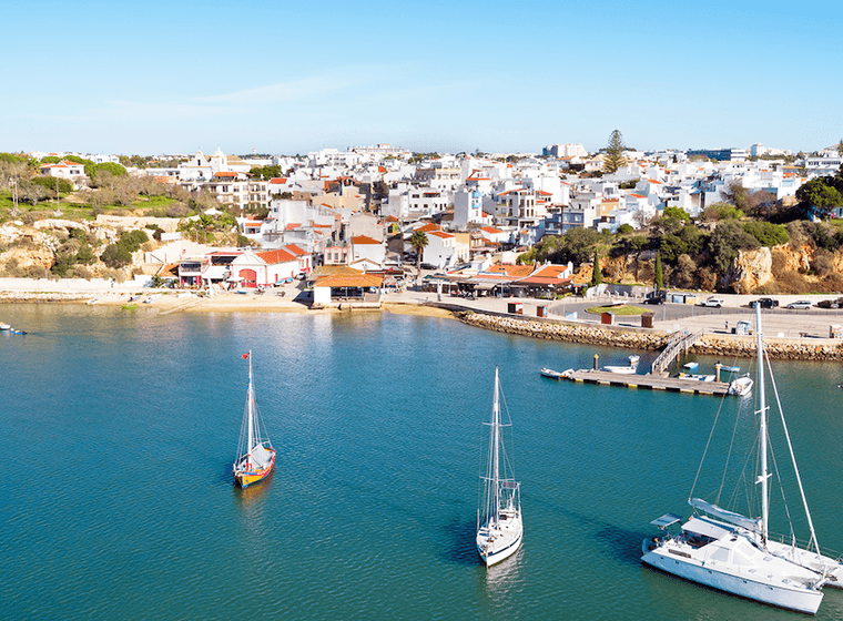 Malerischer Blick auf eine Küstenstadt mit weißen Gebäuden, Segelbooten auf ruhigem Wasser und einem klaren blauen Himmel.