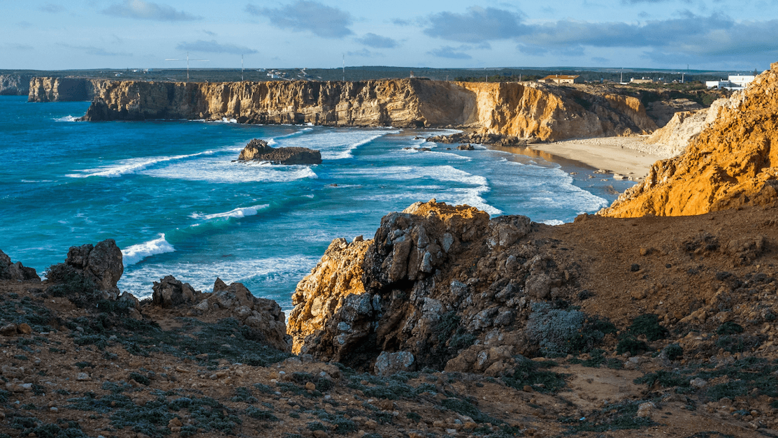 Rocky coastline with cliffs and a sandy beach, waves crashing against the shore under a partly cloudy sky, and sunlight illuminating the landscape.