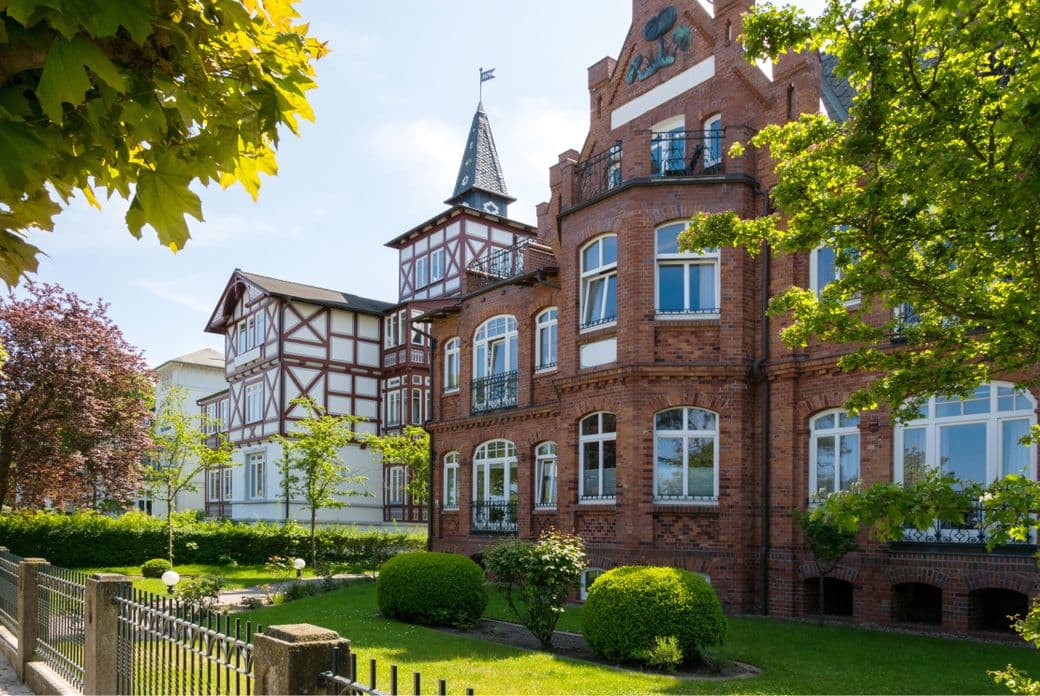 Historic brick and timber-framed buildings with gabled roofs, surrounded by lush greenery and trees under a clear blue sky.