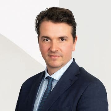 A man in a suit and tie with short dark hair poses against a light background, looking directly at the camera.