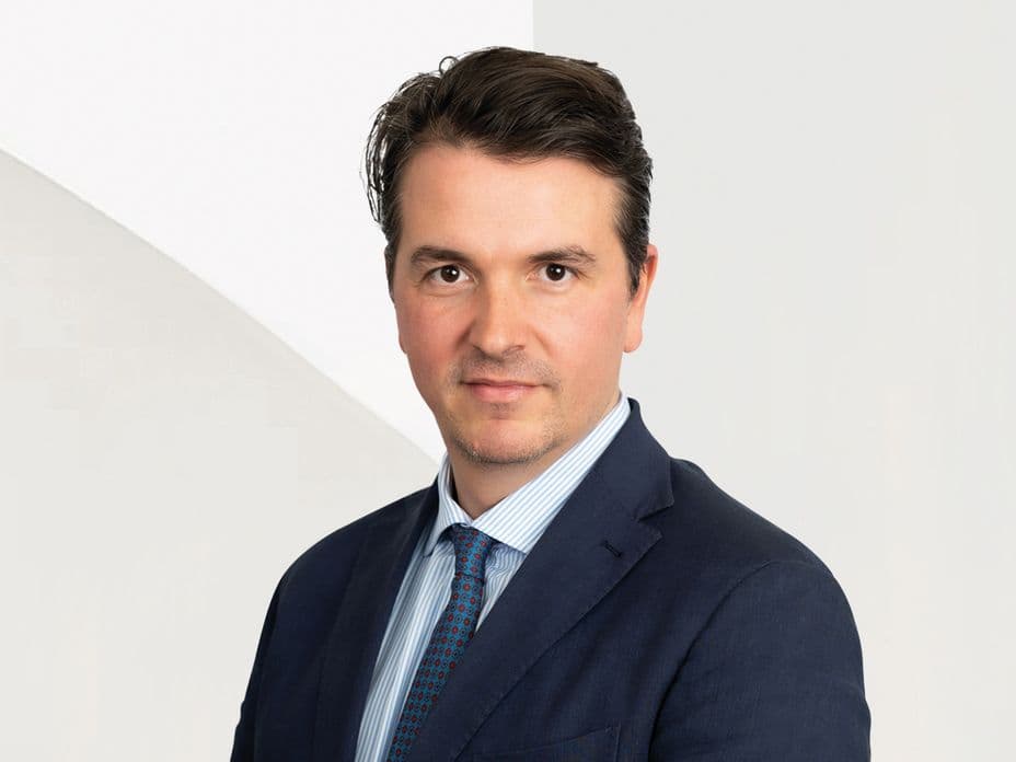 A man in a suit and tie with short dark hair poses against a light background, looking directly at the camera.