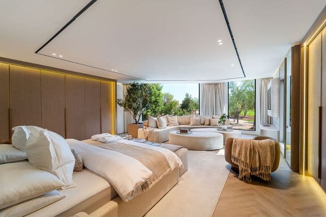 Spacious, modern bedroom with large windows, a round sofa, plants, and warm lighting. Neutral tones and wooden accents create a cozy atmosphere.