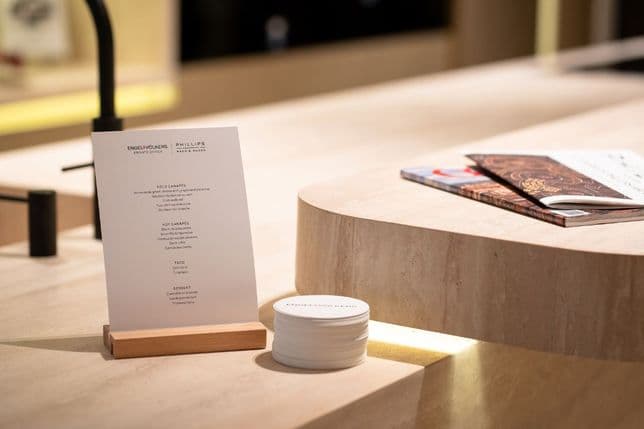 A menu on a wooden stand next to a stack of coasters on a marble countertop, with magazines in the background.