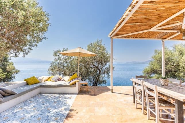 Sunlit outdoor terrace with a built-in lounge area, patterned tiles, a wooden dining table under a bamboo pergola, and panoramic sea views.