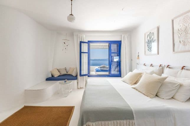 Bright bedroom with white interiors, a large bed, a built-in sofa, and blue window frames opening to a sea view.