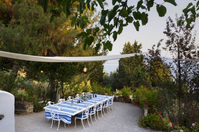 Shaded outdoor dining area with a long table, blue and white striped tablecloth, and surrounding greenery.