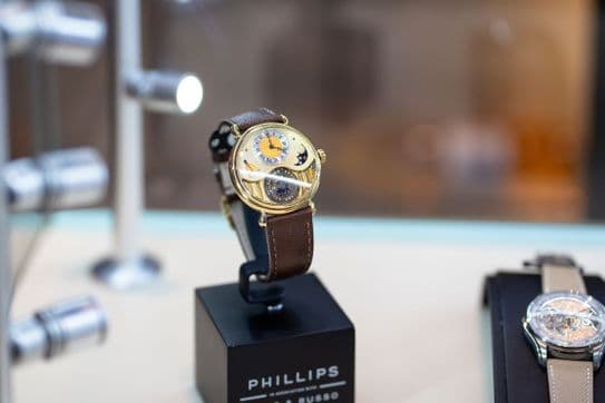 A luxury gold watch with a brown leather strap on display, featuring a moon phase and intricate dial, placed on a black stand.