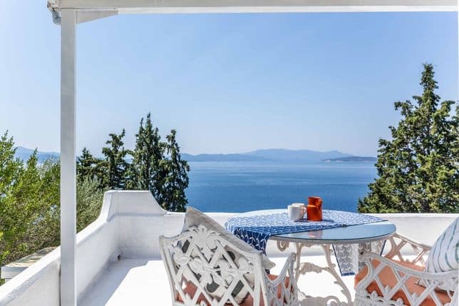 White balcony overlooking the sea with a glass-topped table, patterned tablecloth, and two ornate white chairs with orange cushions.