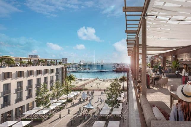 Elegant rooftop lounge and restaurant overlooking Mina Rashid marina, with views of luxury yachts, a sandy promenade, shaded seating areas, and a central plaza featuring a modern sculpture. The scene is lively with people dining and walking under a bright blue sky.