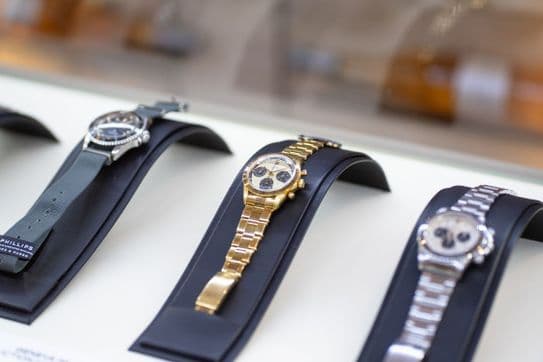 Three luxury watches displayed on black stands, featuring silver, gold, and silver bands with detailed dials, in a well-lit showcase.