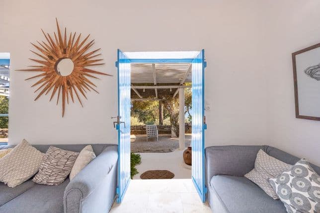 Bright sitting area featuring grey sofas, a sunburst mirror, and open bright blue double doors leading to a shaded outdoor patio.