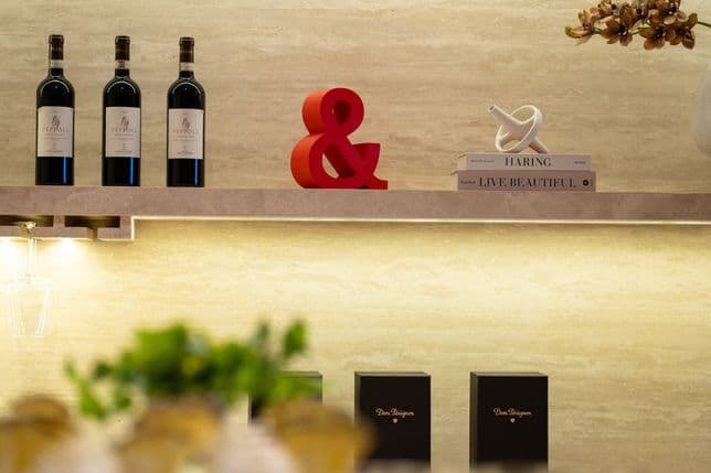 Shelf with wine bottles, a red ampersand, books, and decor against a beige wall. Below, blurred greenery and black boxes are visible.