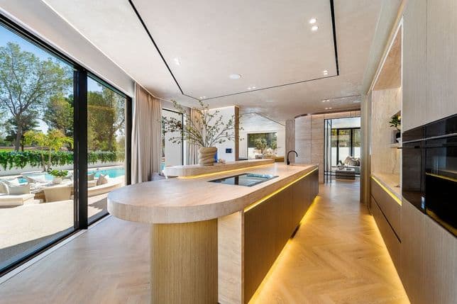 Modern kitchen with a large island, sleek wooden cabinetry, and floor-to-ceiling windows overlooking a patio with outdoor seating.