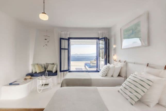 Bright bedroom with white interiors, twin beds, a built-in sofa, and blue window frames opening to a sea view.