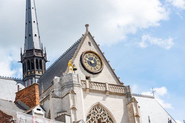 Exterior view of St. Rumbold's Cathedral in Mechelen, Belgium, featuring a clock, spire, and golden statue against a blue sky.