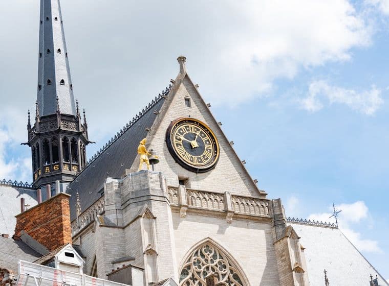 Buitenaanzicht van de Sint-Romboutskathedraal in Mechelen, België, met een klok, torenspits en gouden beeld tegen een blauwe lucht.