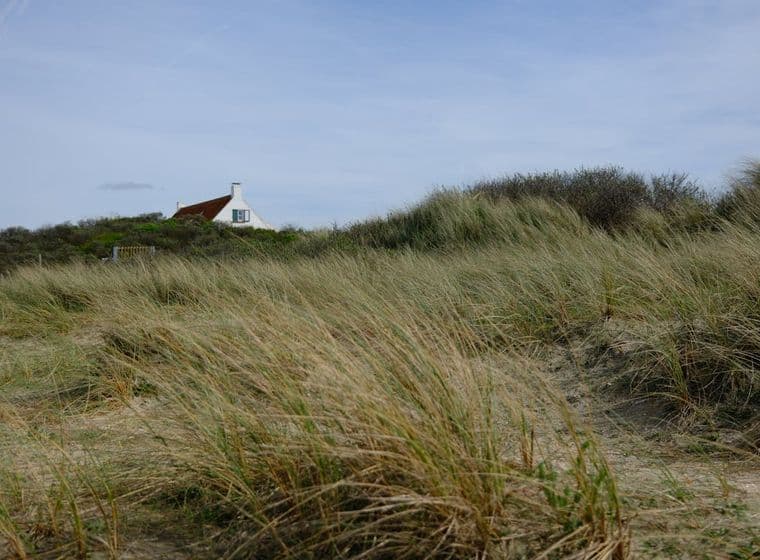 Strandhuis met rood dak, gezien vanaf de met gras begroeide duinen onder een blauwe hemel.