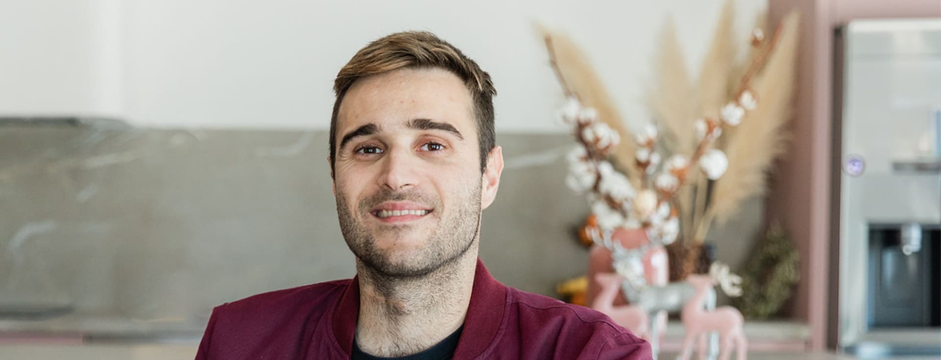 A smiling man with short brown hair and a burgundy jacket stands in a modern kitchen.