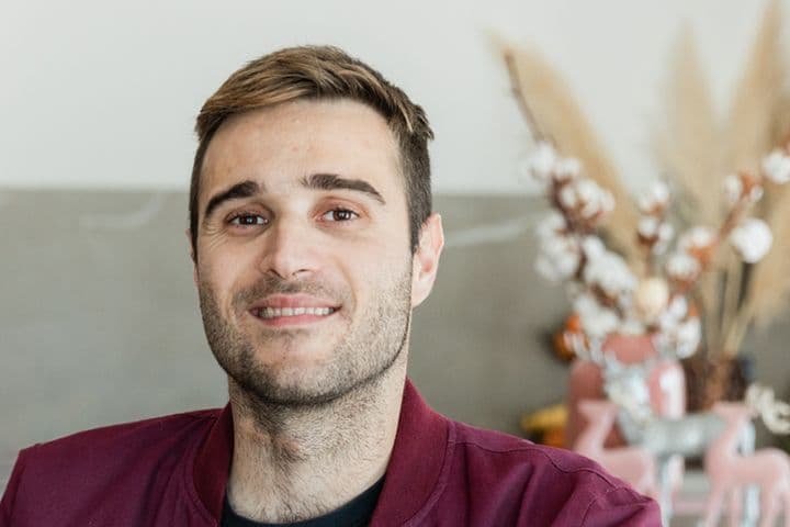 A smiling man with short brown hair and a burgundy jacket stands in a modern kitchen.