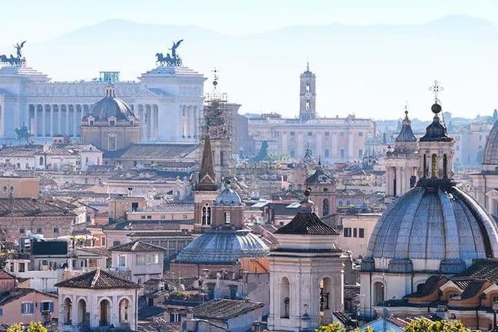 Panorama di Roma con cupole, tetti e il Monumento a Vittorio Emanuele II sotto un cielo blu.