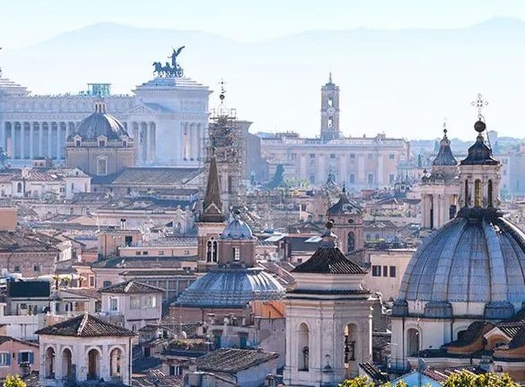 Panorama di Roma con cupole, tetti e il Monumento a Vittorio Emanuele II sotto un cielo blu.
