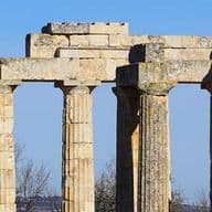 Ruinen des Tempels der Aphaia auf Ägina, Griechenland. Steinsäulen erheben sich vor einem klaren, blauen Himmel. Kahle Bäume sind links zu sehen.