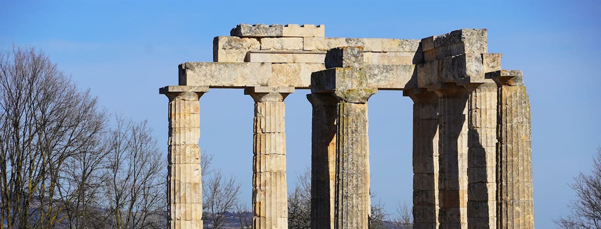 Ruins of the Temple of Aphaia on Aegina, Greece. Stone columns stand against a clear blue sky. Bare trees are visible on the left.