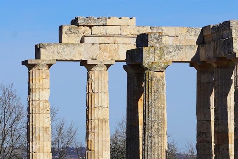 Ruinen des Tempels der Aphaia auf Ägina, Griechenland. Steinsäulen erheben sich vor einem klaren, blauen Himmel. Kahle Bäume sind links zu sehen.