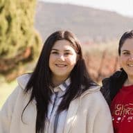 Two women, the proud owners of Palyvou Winery, are smiling outdoors. They stand side by side in a scenic vineyard setting with greenery and hills in the background. Both are wearing jackets, and one has a shirt with white text.
