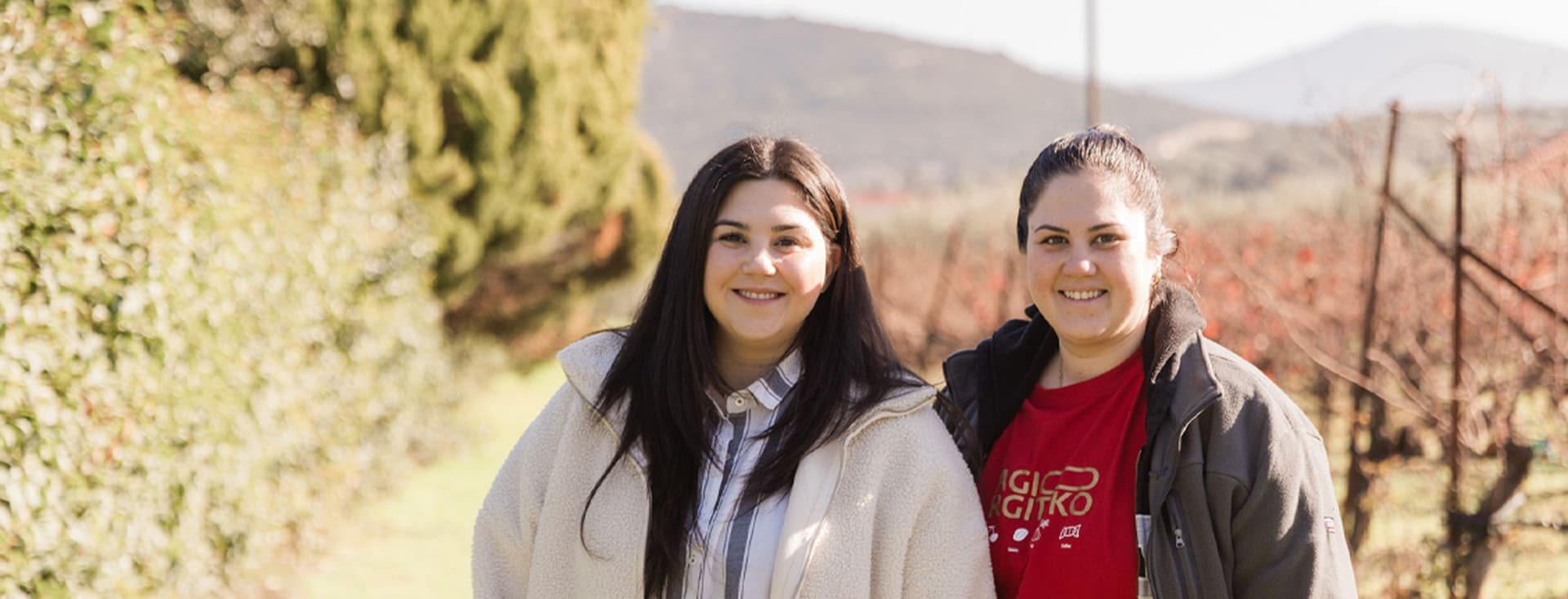 Two women, the proud owners of Palyvou Winery, are smiling outdoors. They stand side by side in a scenic vineyard setting with greenery and hills in the background. Both are wearing jackets, and one has a shirt with white text.