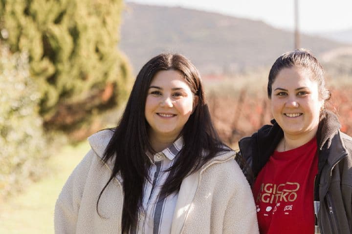 Two women, the proud owners of Palyvou Winery, are smiling outdoors. They stand side by side in a scenic vineyard setting with greenery and hills in the background. Both are wearing jackets, and one has a shirt with white text.