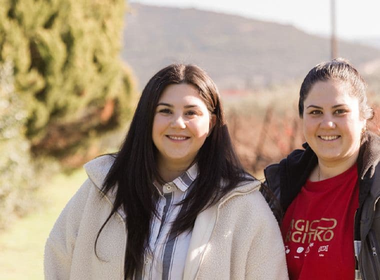 Two women, the proud owners of Palyvou Winery, are smiling outdoors. They stand side by side in a scenic vineyard setting with greenery and hills in the background. Both are wearing jackets, and one has a shirt with white text.