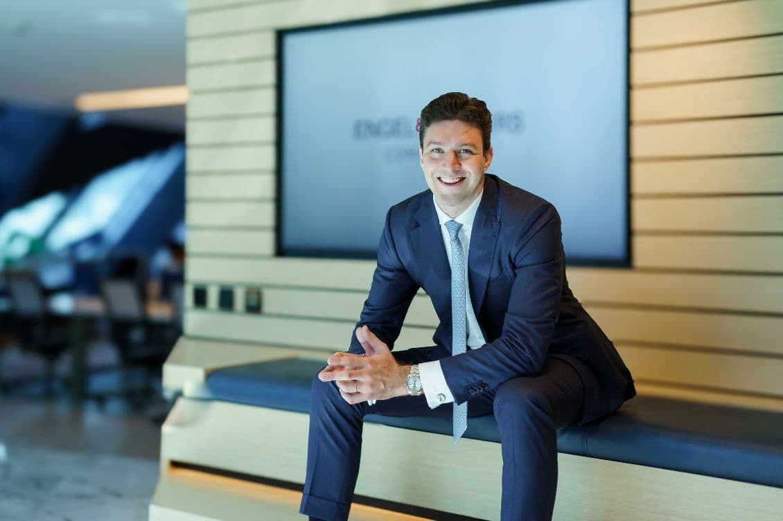 Rogier Arends in a suit sits on a bench in a modern office with a smile, hands clasped. A large screen is visible in the background.