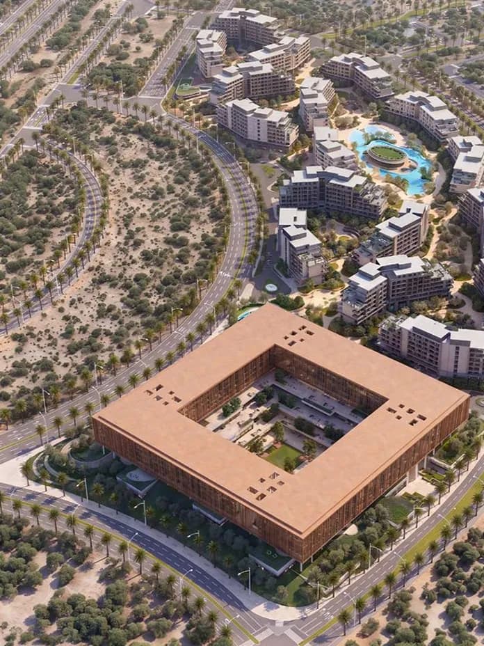 An aerial view of a desert development: a large square brown building with a central courtyard, surrounded by curved roads and modern residential blocks with a pool.