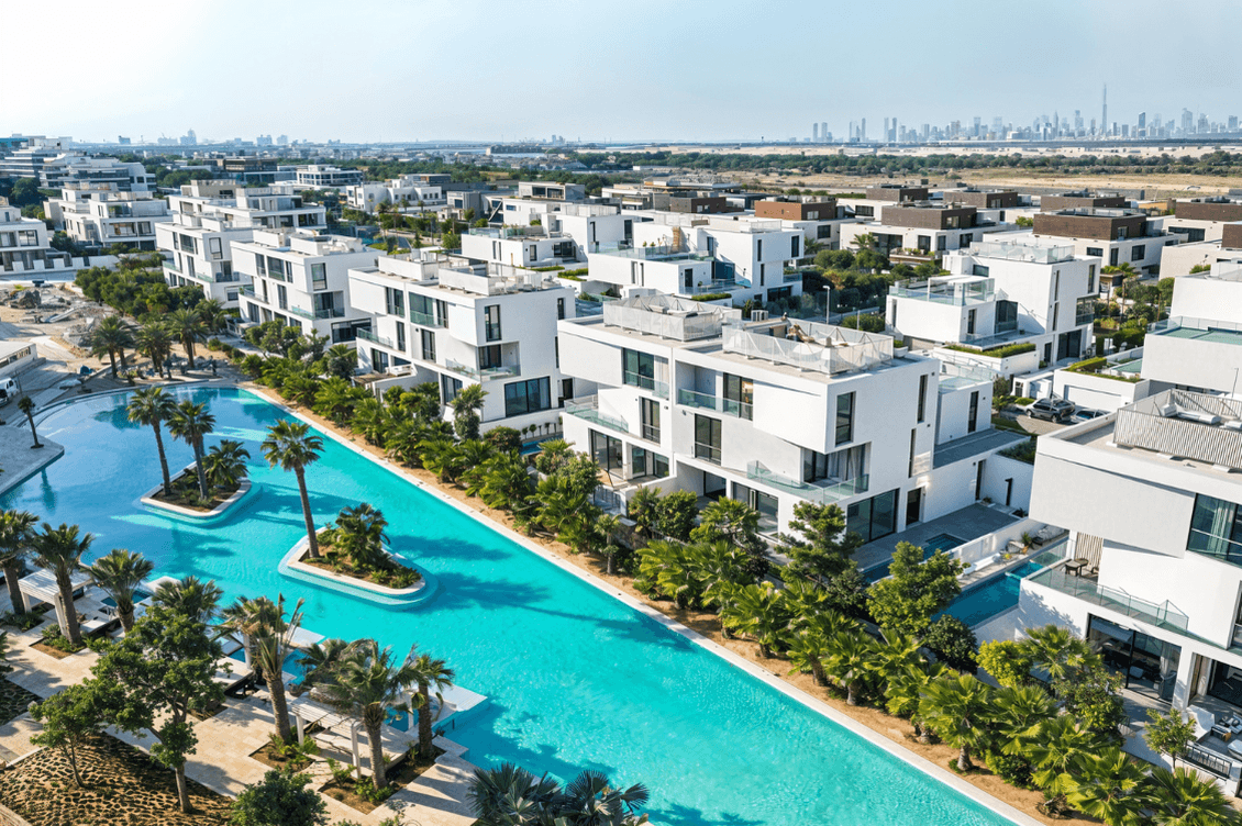 Aerial view of modern white buildings with balconies, surrounded by palm trees and a long turquoise pool, with a city skyline in the distance.