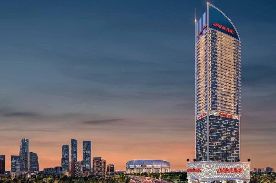 Nachtansicht des ikonischen Danube ASPIRZ-Turms, beleuchtet in der Dubai Sports City von Danube Properties, mit umliegender Stadtsilhouette und Straßen.