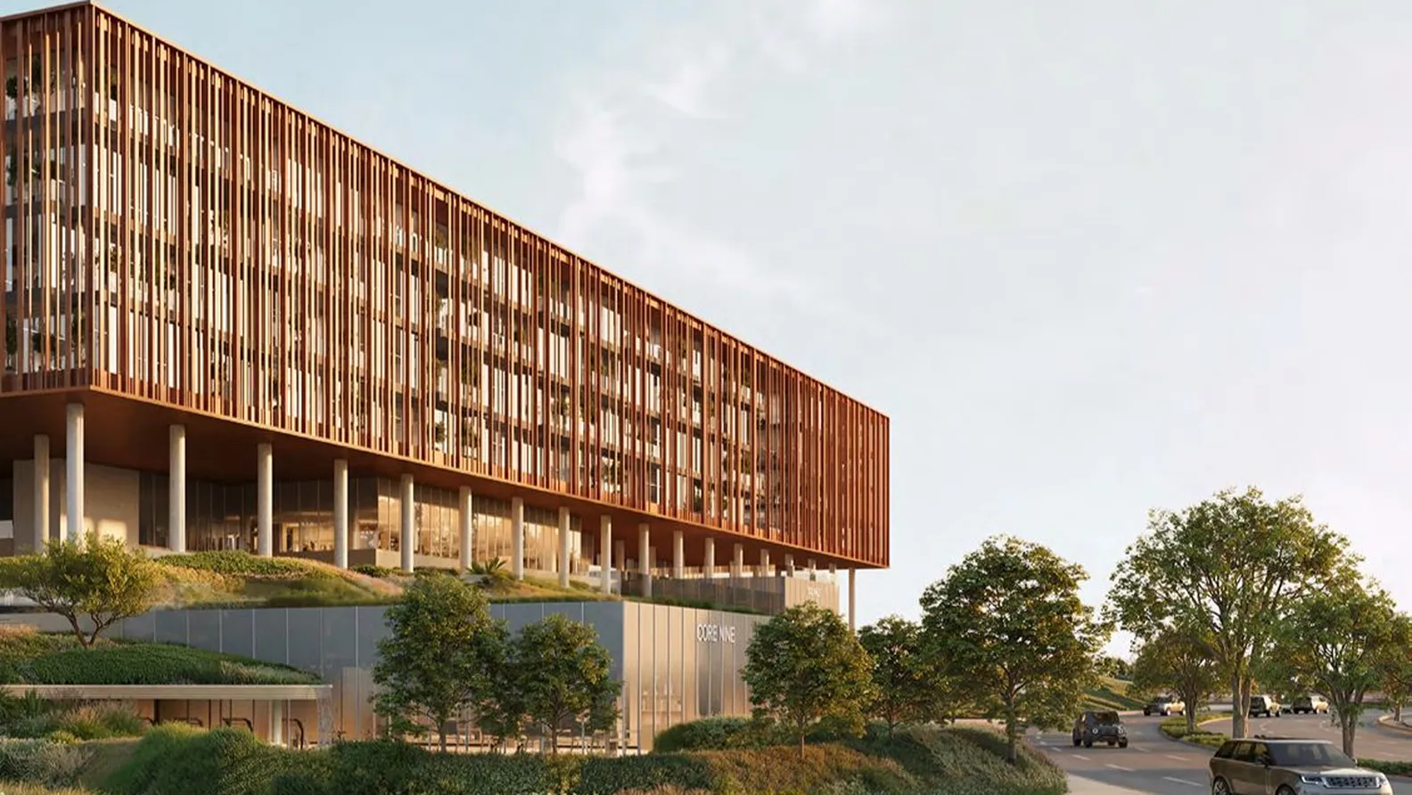 Contemporary glass-fronted building with a vertical wooden slat façade, elevated on columns, surrounded by trees and a curved road with cars.