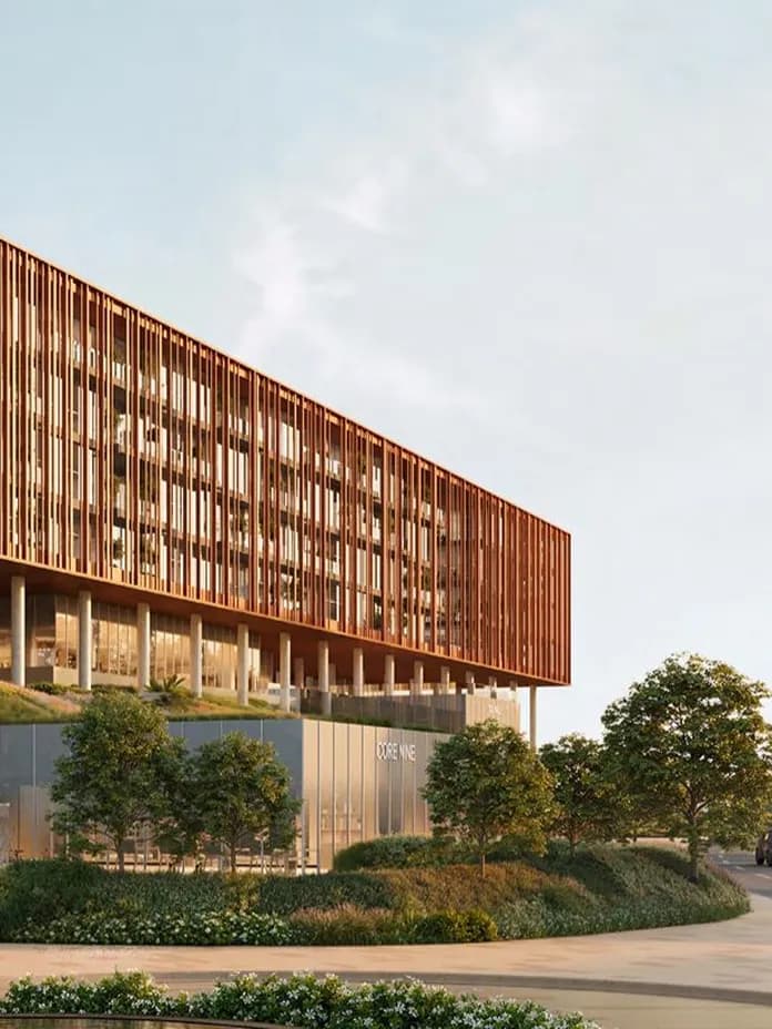 Contemporary glass-fronted building with a vertical wooden slat façade, elevated on columns, surrounded by trees and a curved road with cars.