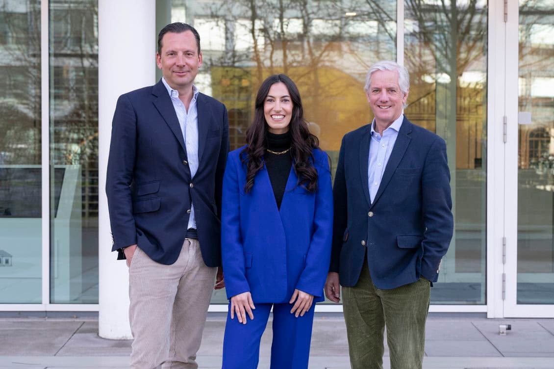 Three people in business attire stand smiling outside a glass building entrance.